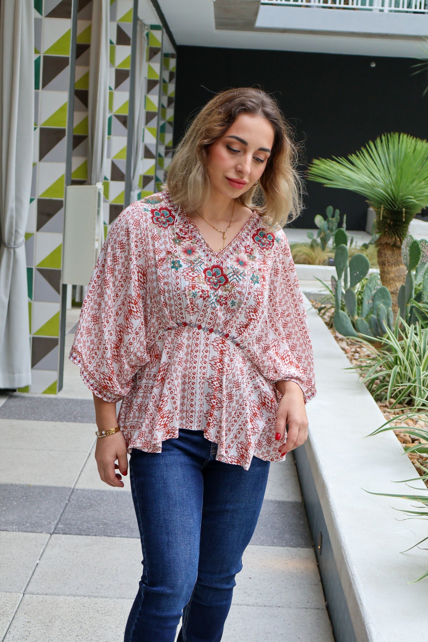 Woman in a floral blouse and blue jeans standing in an outdoor setting with plants and geometric wall patterns.