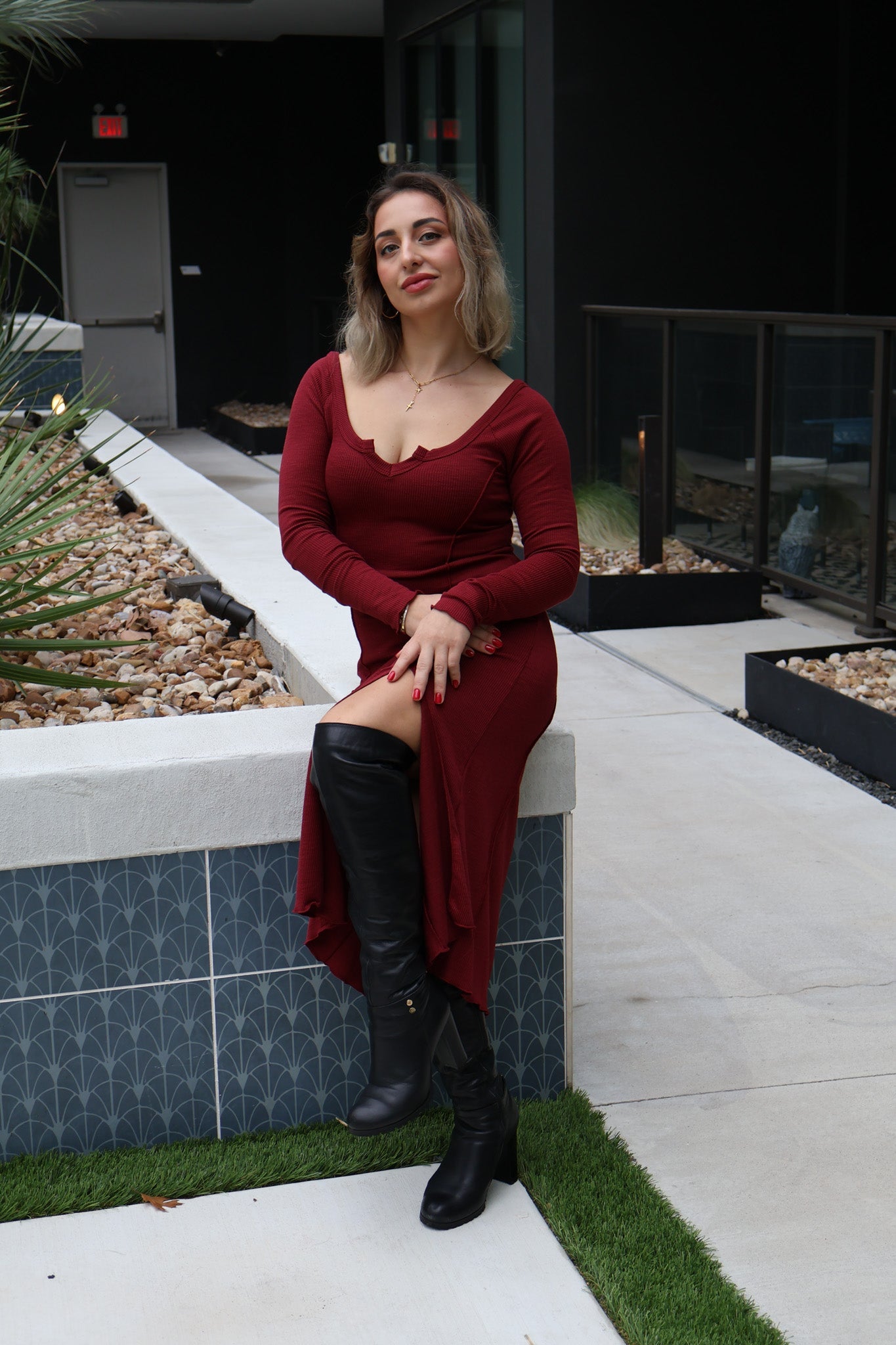 Woman in a red dress and black knee-high boots sitting on a ledge outdoors.