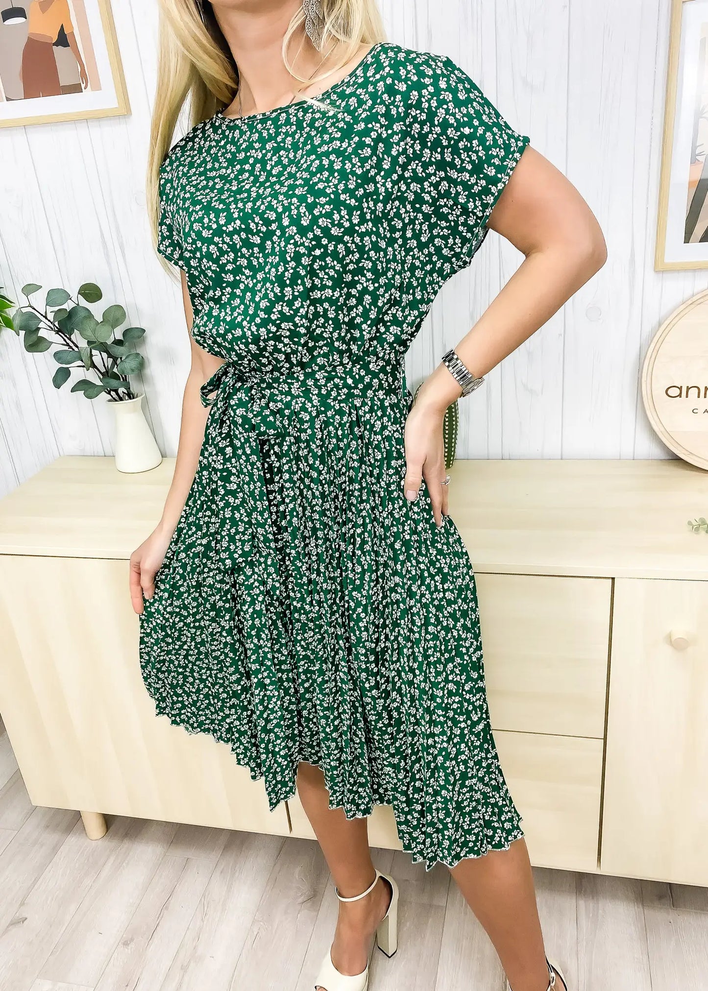 A model wearing a green dress featuring a round neck line, short sleeves, tie-waist detail, pleated skirt, and below knee length. Pair it with a necklace and boots for an elegant wine tasting in San Luis Obispo look.