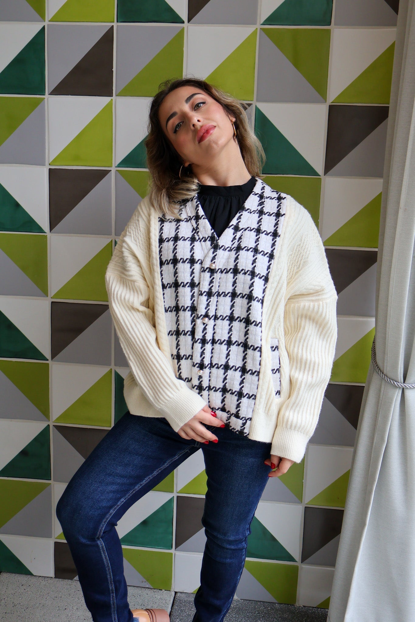 Woman wearing a cream cardigan over a patterned shirt against a geometric tile wall.
