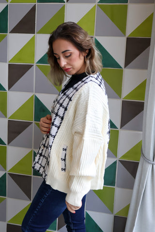 Woman wearing a white sweater with a patterned scarf against a geometric tile wall.