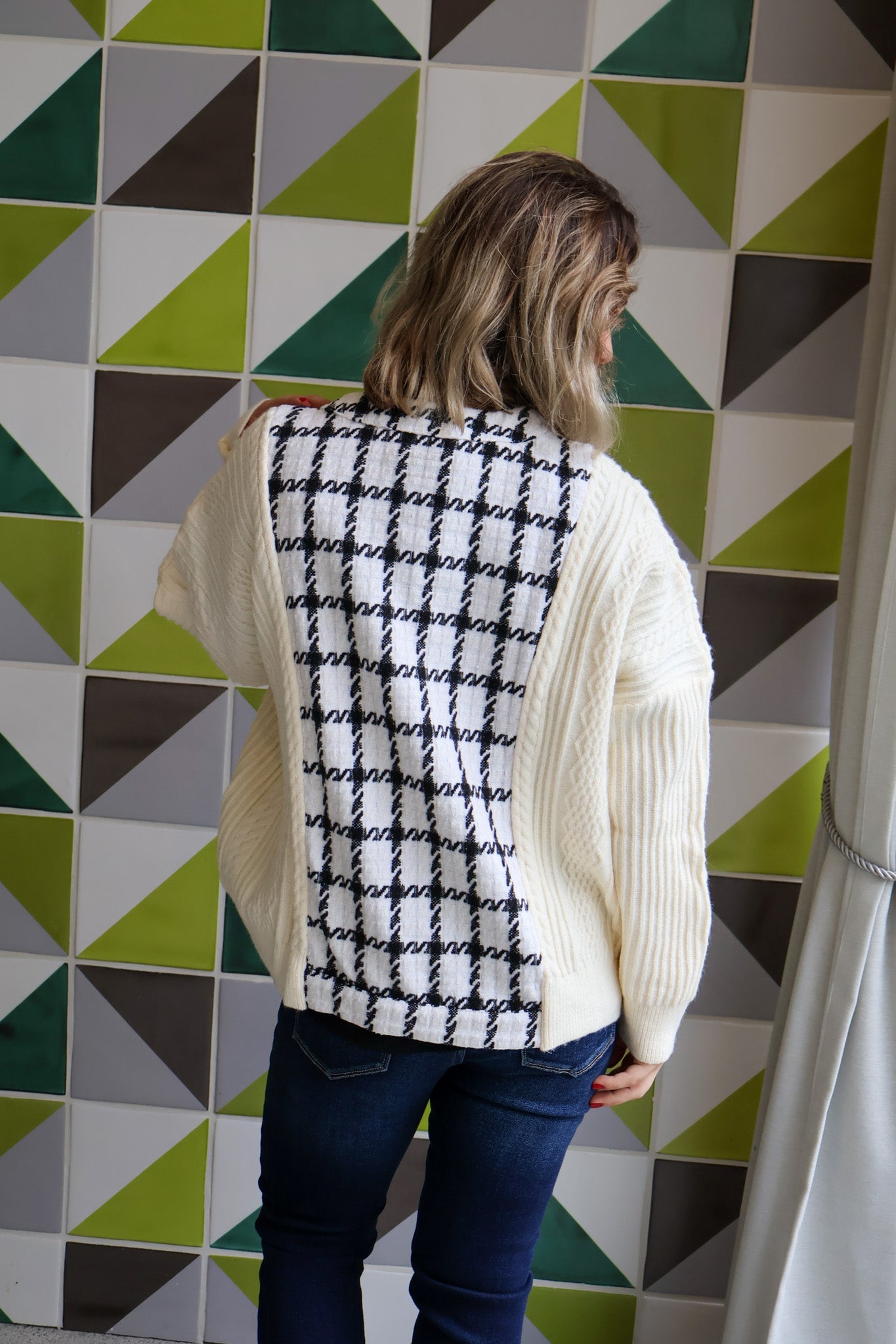 Person wearing a white cardigan over a black and white patterned shirt against a geometric tile wall.