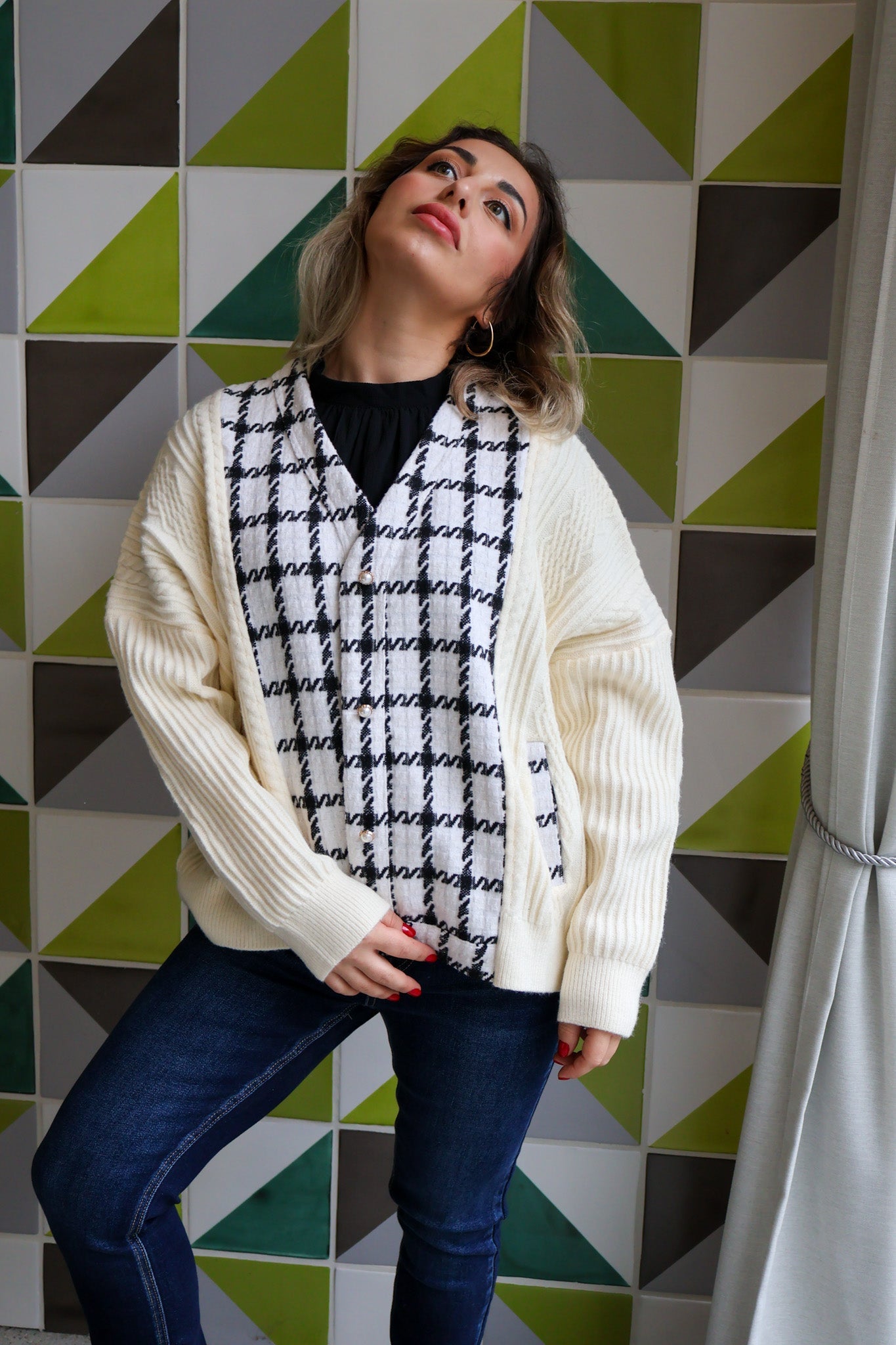 Woman wearing a white cardigan over a black and white patterned scarf with a geometrically patterned wall in the background.