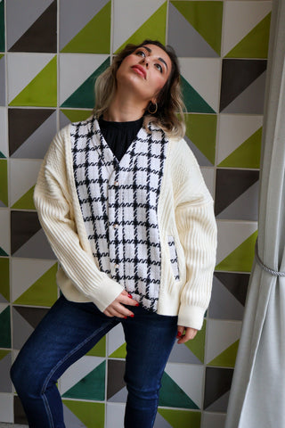 Woman wearing a white cardigan over a black and white patterned scarf with a geometrically patterned wall in the background.