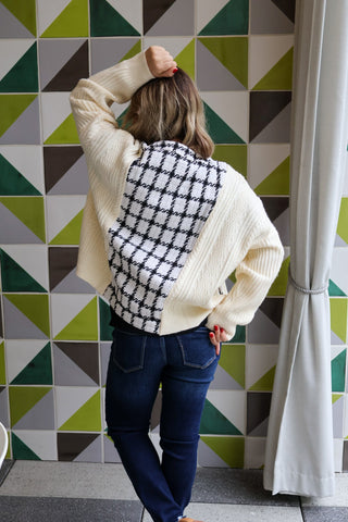 Person wearing a white sweater with a black and white patterned back, standing against a geometric tile wall.
