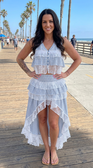 A woman stands on a sunny wooden boardwalk wearing a two-piece gingham and lace set consisting of a sleeveless crop top and a high-low tiered skirt.