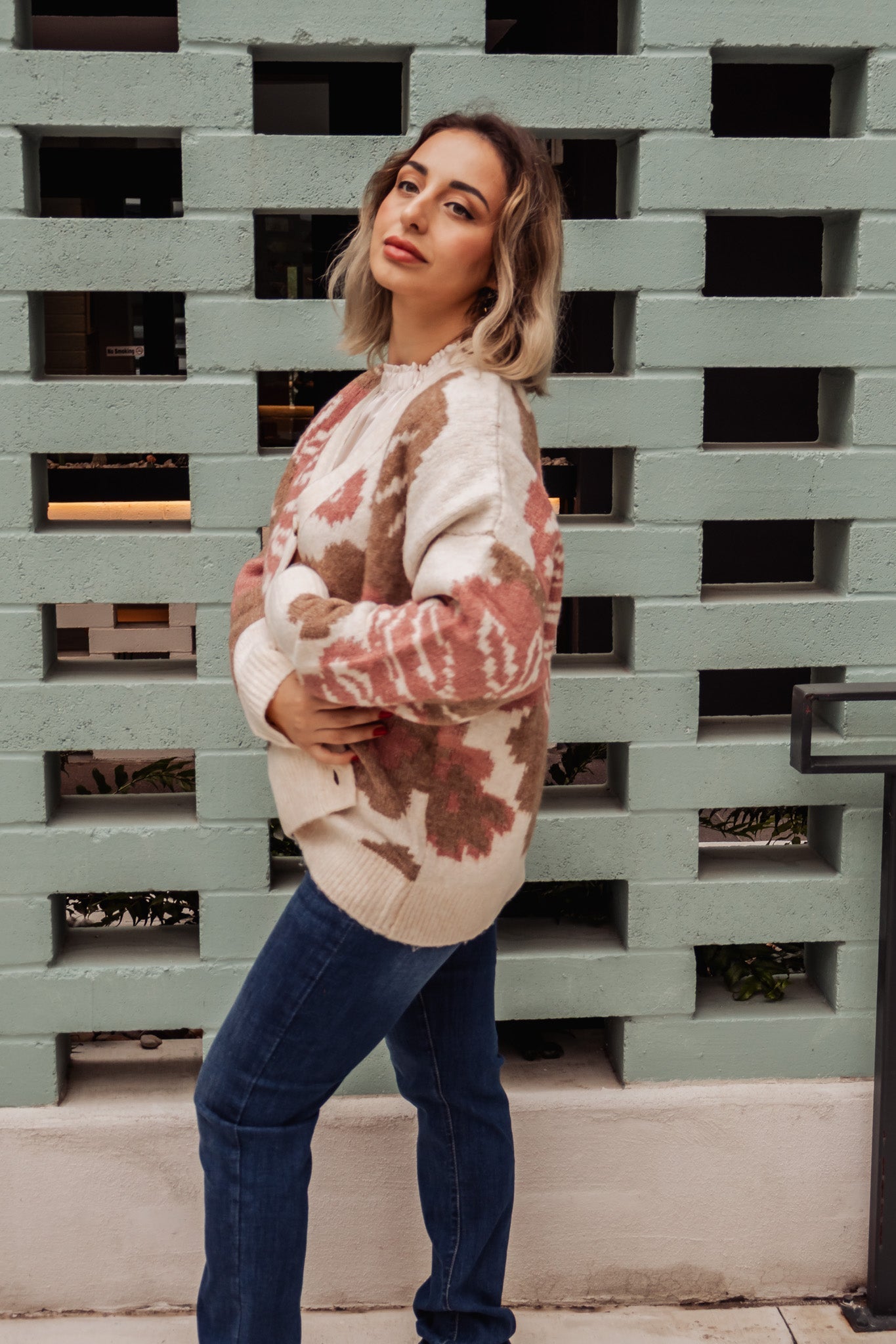 Woman wearing a tie-dye sweater and jeans standing against a textured wall.