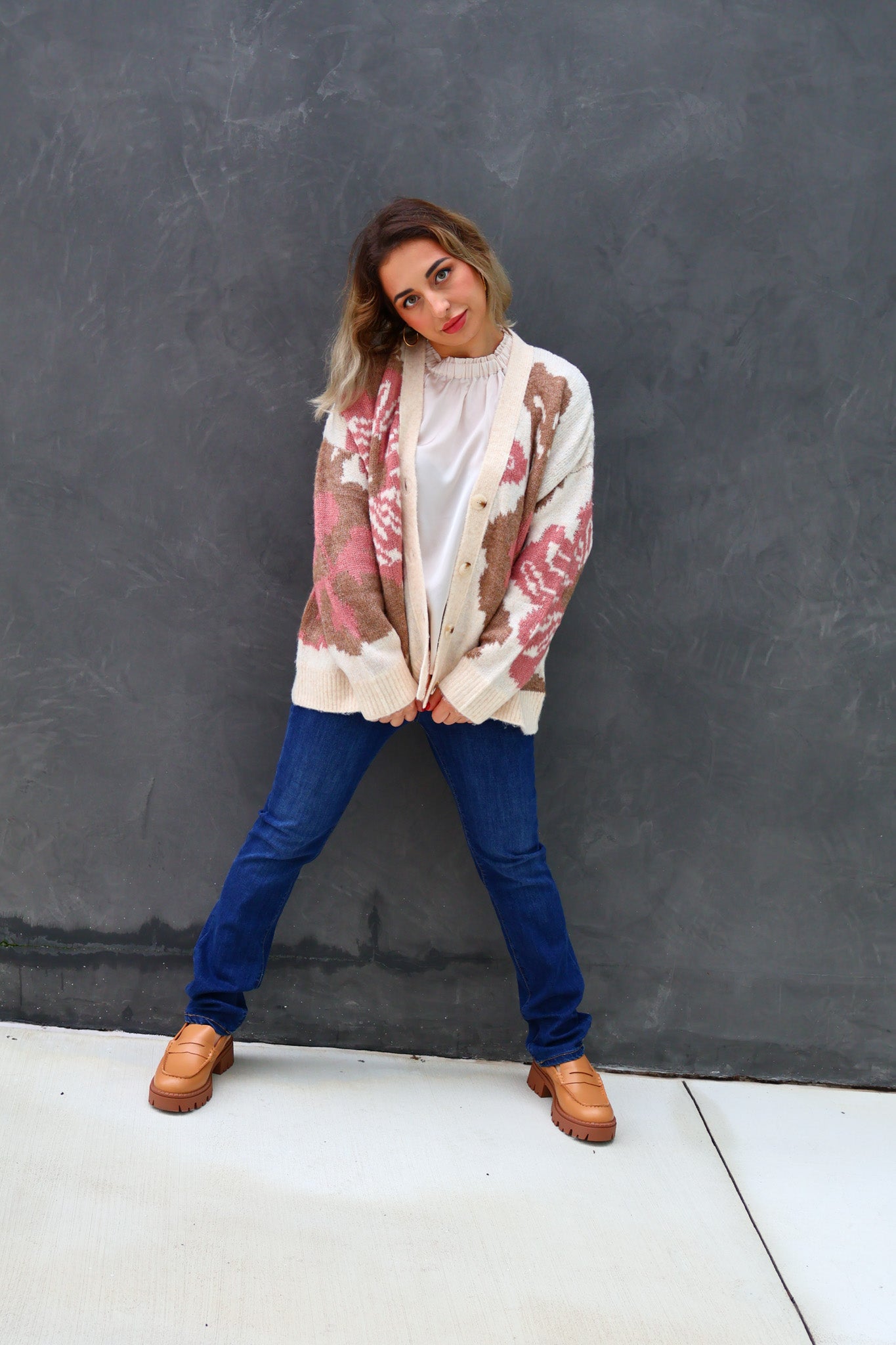 Woman wearing a tie-dye cardigan, white turtleneck, blue jeans, and brown boots against a gray wall.