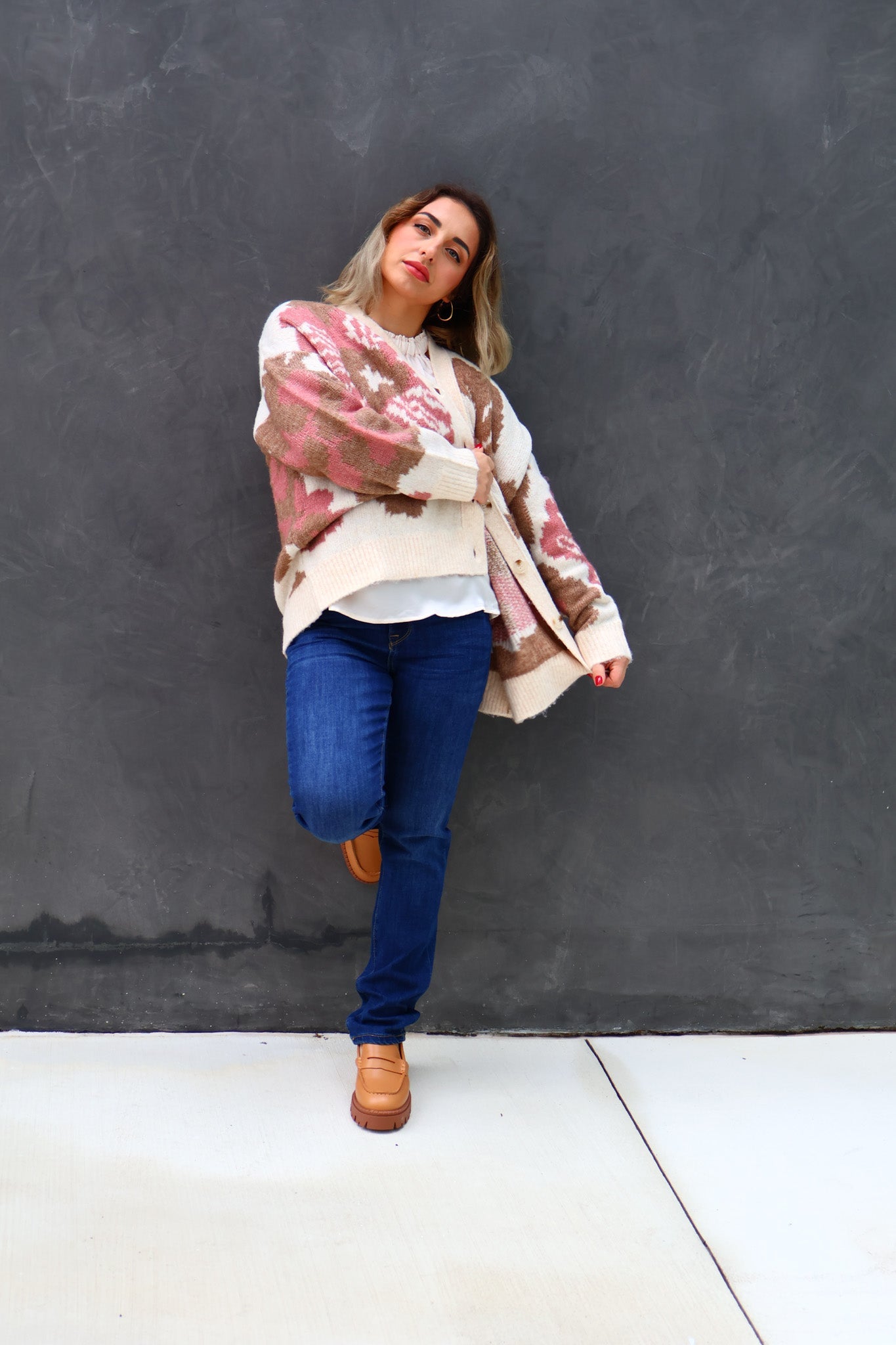 Woman wearing a patterned shawl over a white shirt and blue jeans against a dark gray wall.
