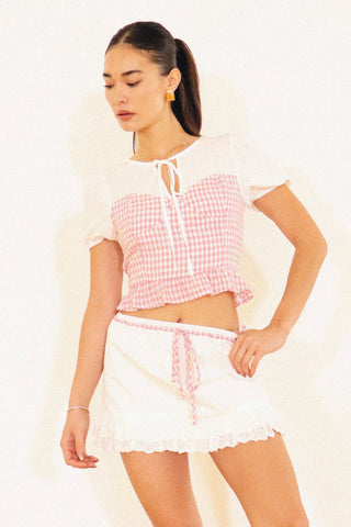 A woman modeling a pink and white gingham crop top with short puff sleeves and a white mini skirt with a matching gingham tie belt, against a plain white background.