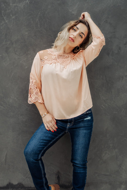 Woman wearing a light pink blouse with lace details and blue jeans against a gray background