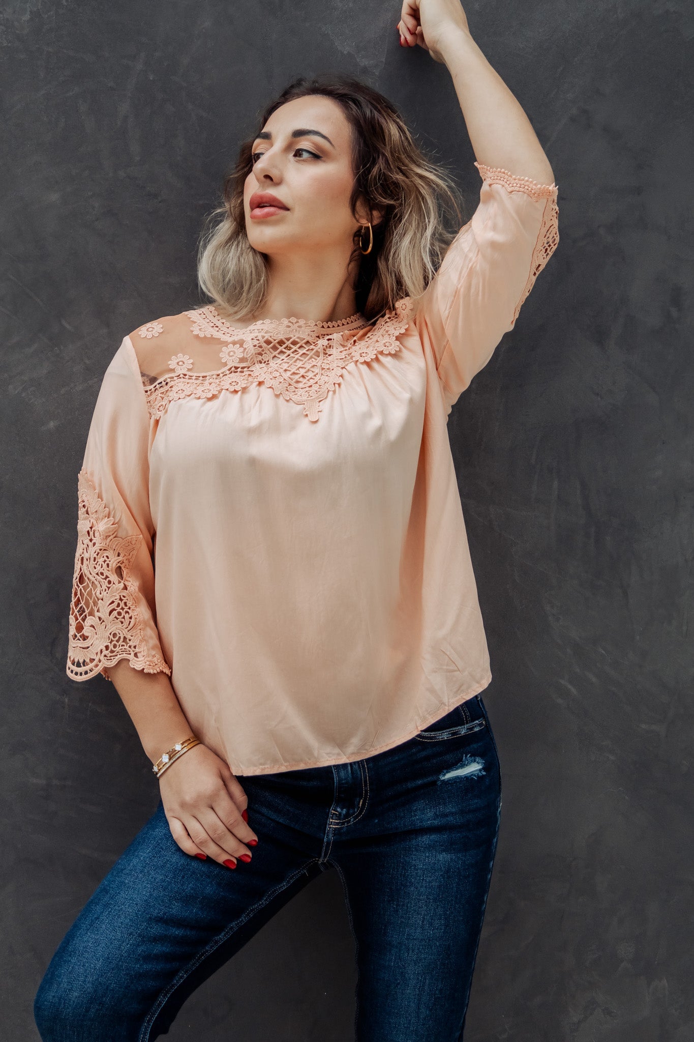 Woman wearing a peach blouse with lace details and blue jeans against a dark background