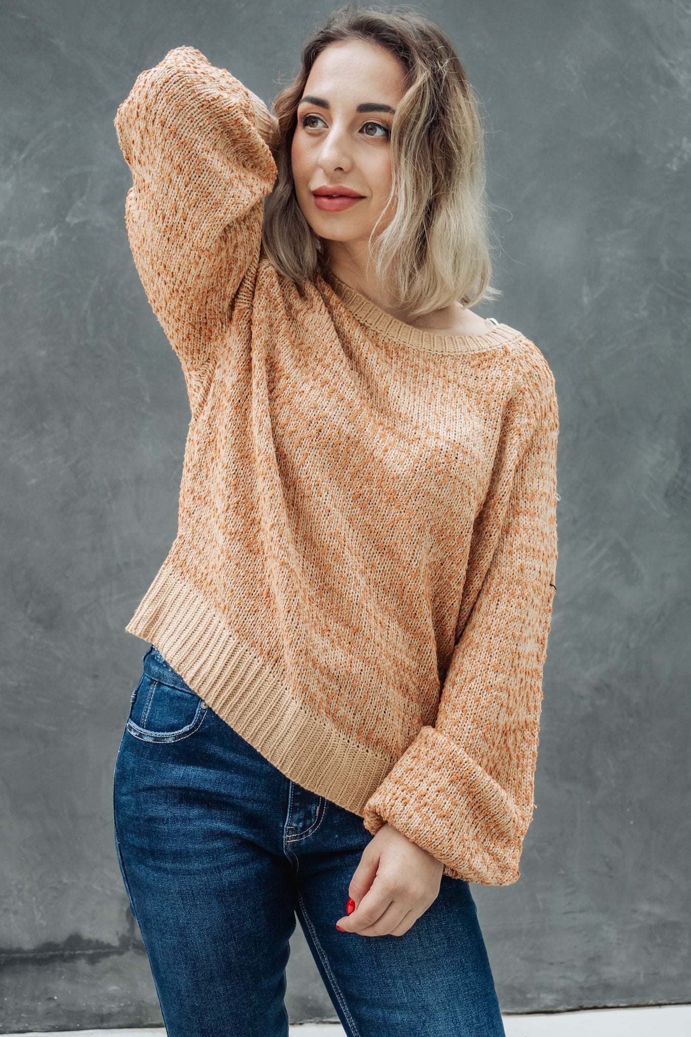 Woman wearing a beige sweater and blue jeans against a gray background
