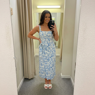 A woman takes a mirror selfie in a fitting room wearing a light blue and white floral print midi dress with a tie waist, paired with white slide sandals.