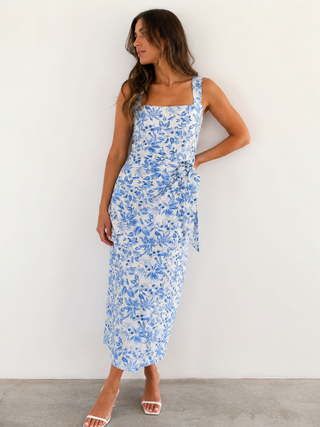 A woman modeling a sleeveless midi dress with a blue floral print on a white background, standing against a plain white wall.