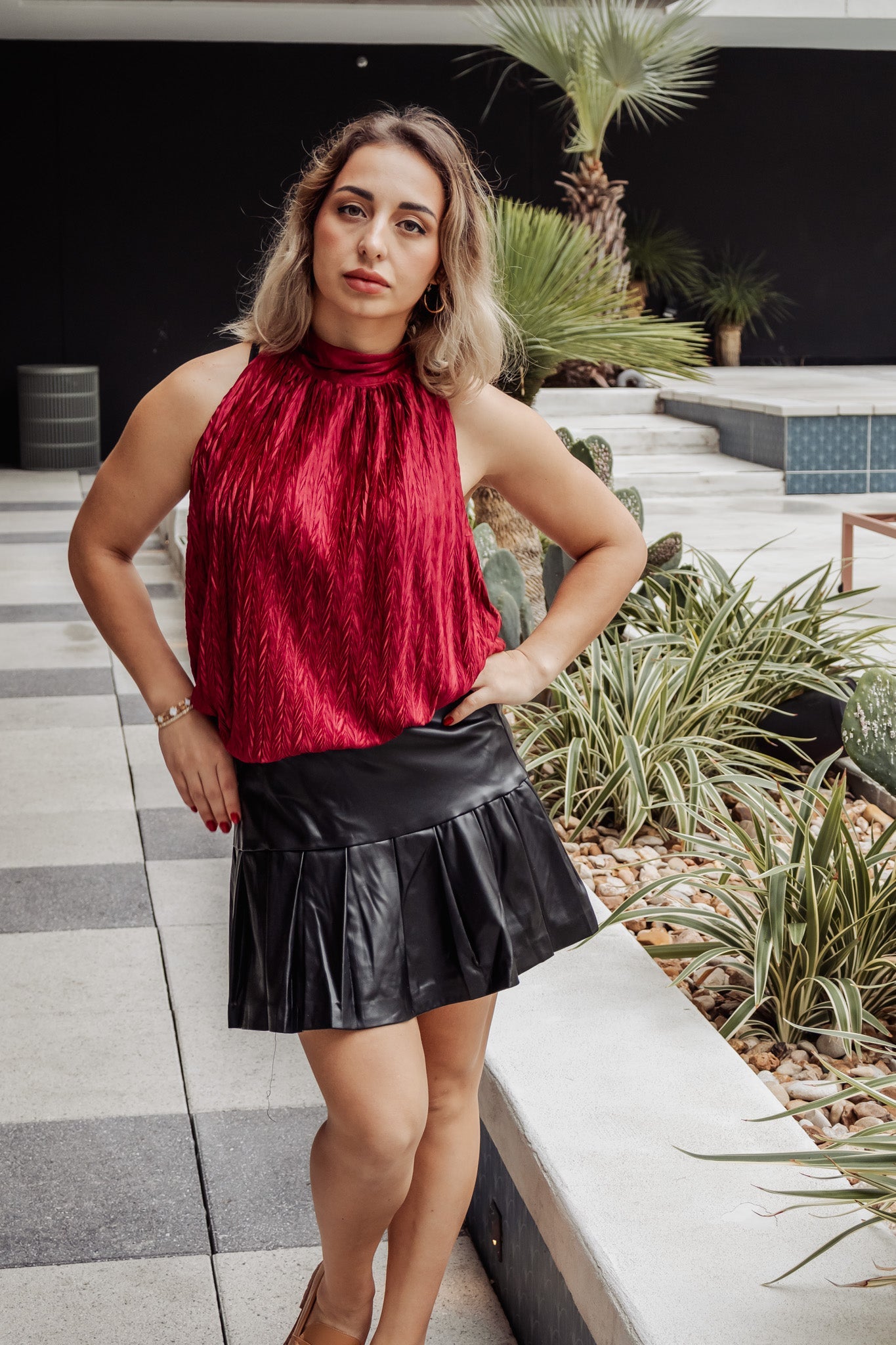 Woman in a red top and black skirt standing outdoors with plants and steps in the background