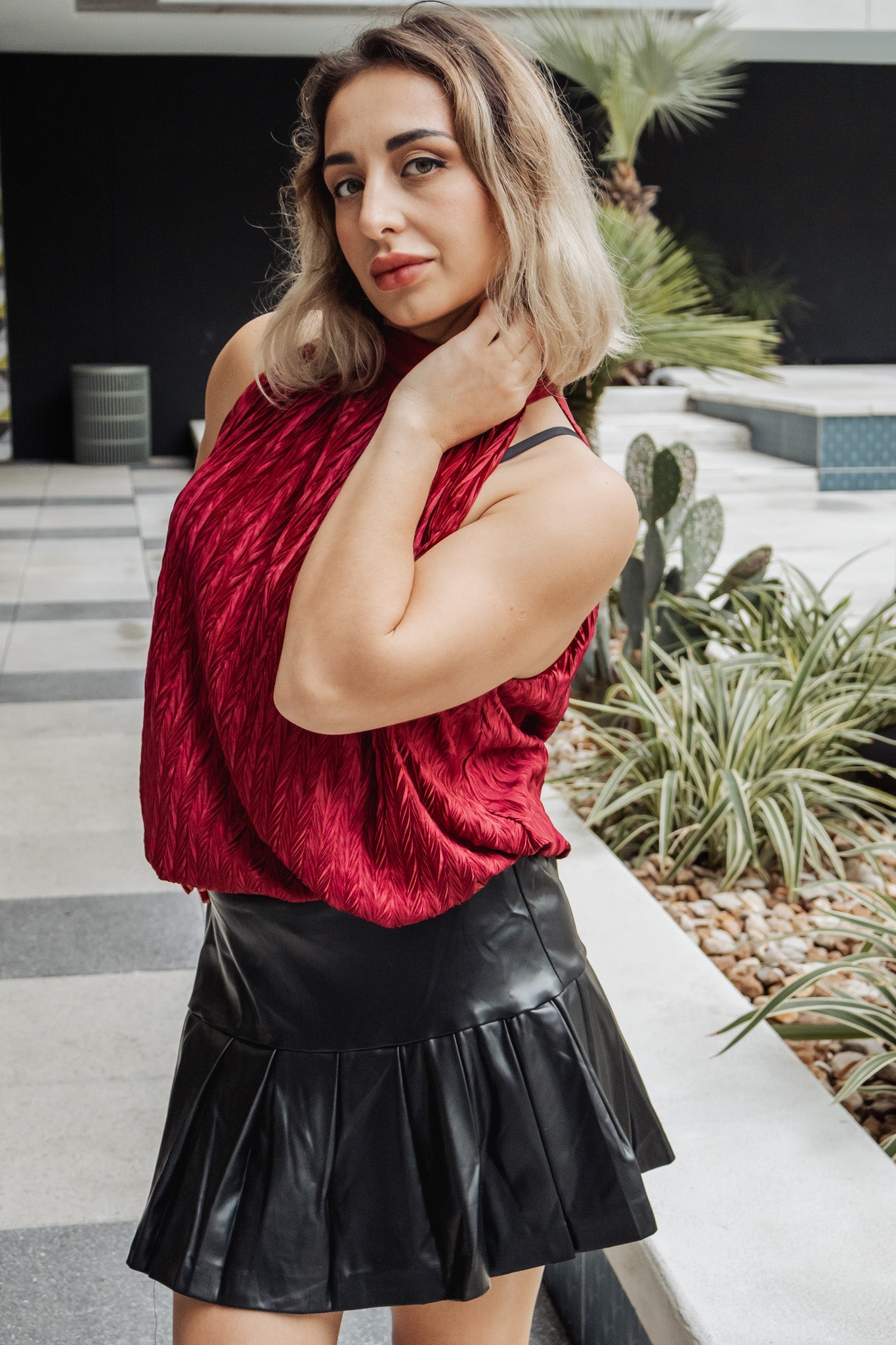 Woman wearing a red top and black leather skirt outdoors with plants in the background