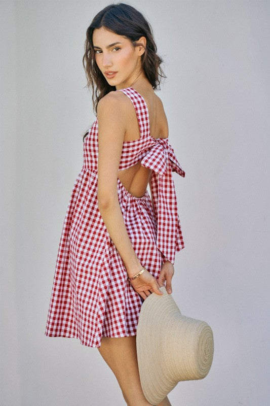 Woman wearing a red and white checkered dress with an open back, holding a straw hat against a plain background.