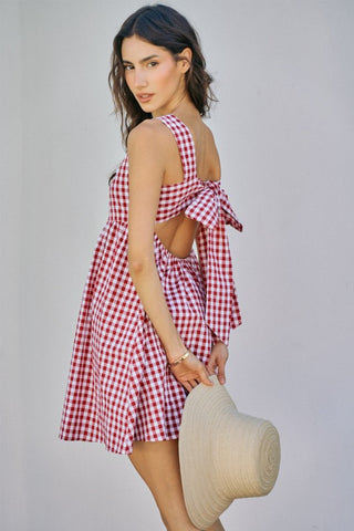Woman wearing a red and white checkered dress with an open back, holding a straw hat against a plain background.