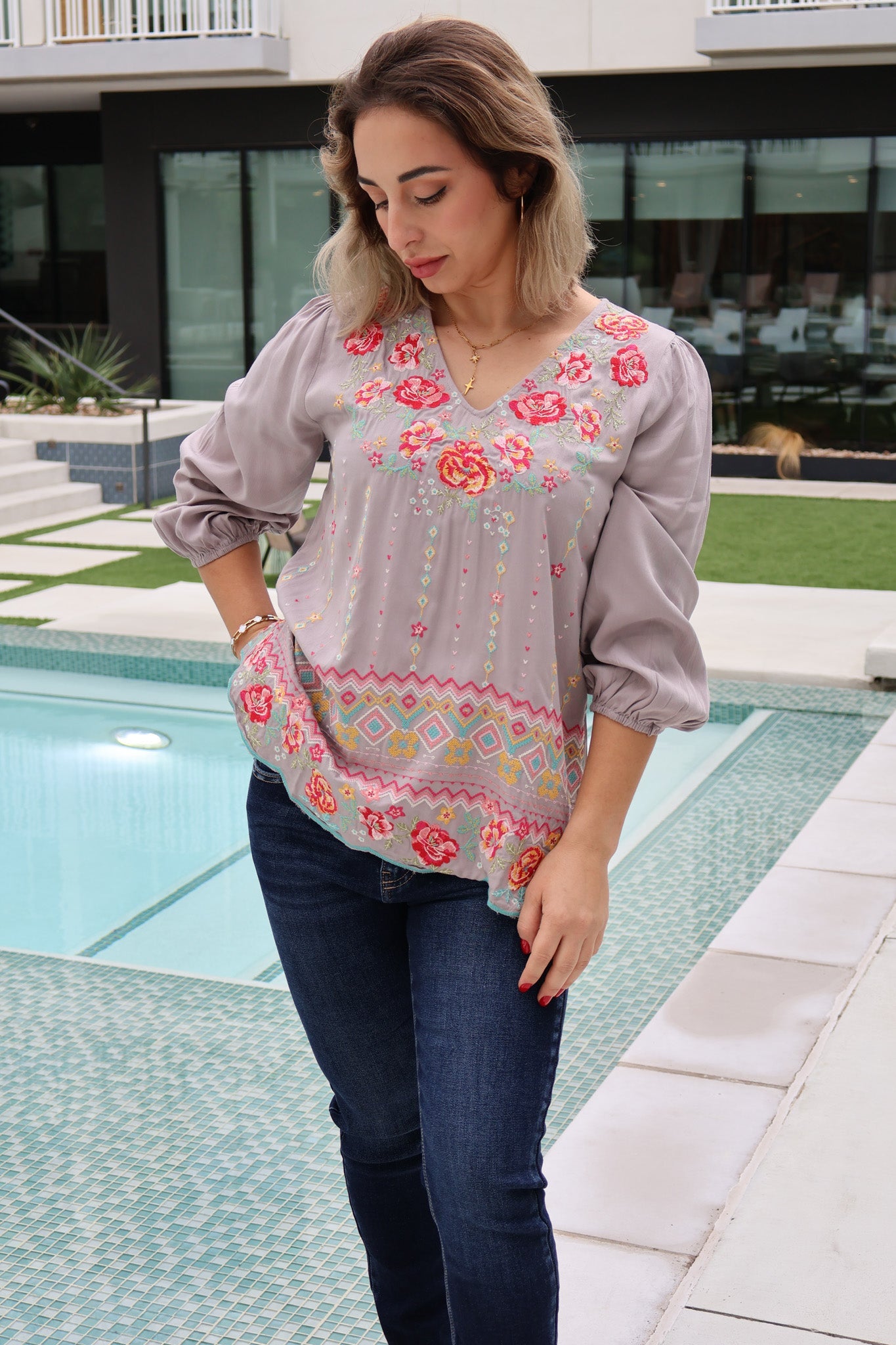 Woman wearing a gray blouse with floral embroidery by a poolside.