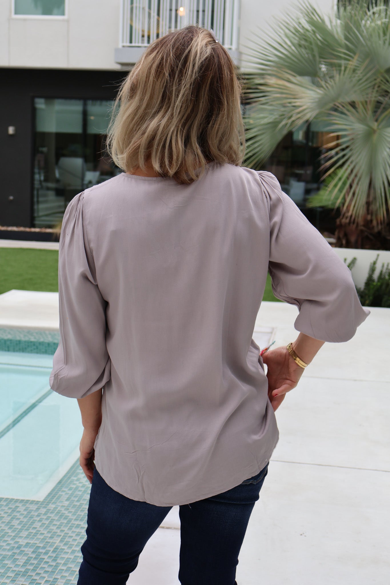 Person wearing a light gray blouse standing by a poolside with a building and palm tree in the background.