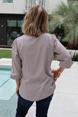 Person wearing a light gray blouse standing by a poolside with a building and palm tree in the background.