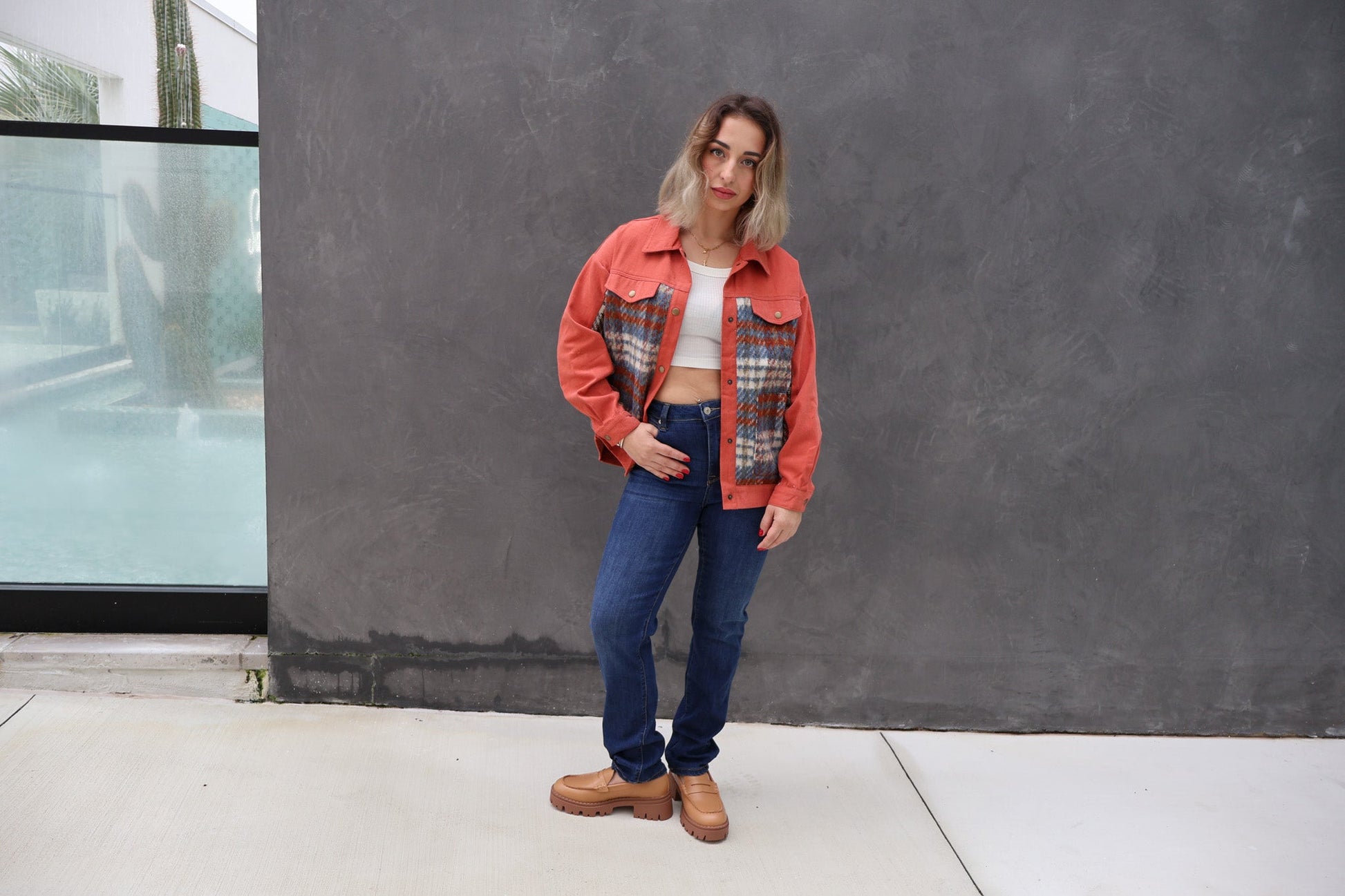 Woman wearing a red plaid jacket and blue jeans standing against a gray wall.