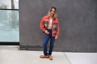 Woman wearing a red plaid jacket and blue jeans standing against a gray wall.