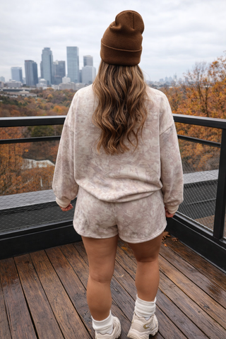 Person wearing a beige sweater and shorts set with a brown beanie, standing on a wooden deck with a cityscape in the background.