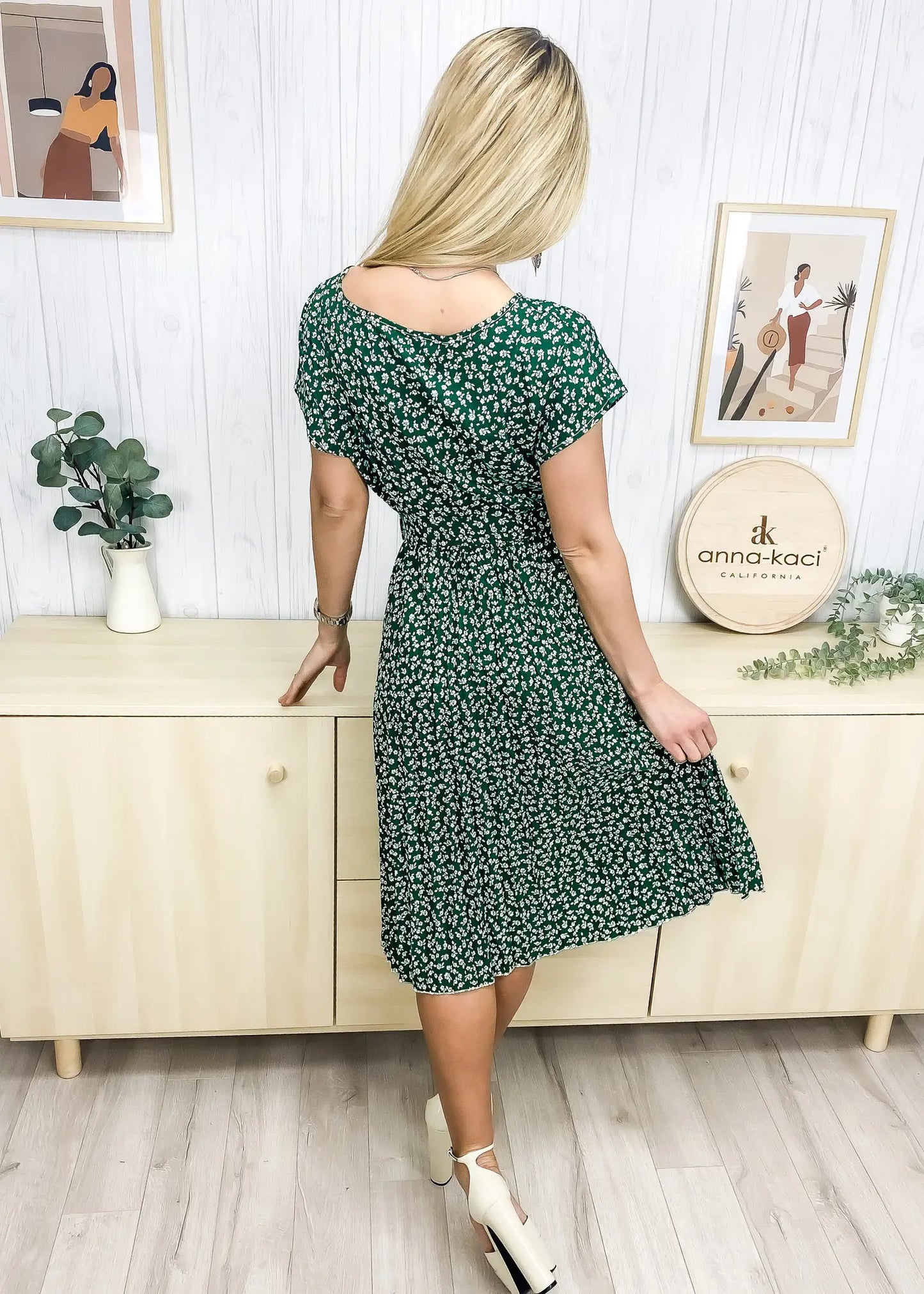 A model wearing a green dress featuring a round neck line, short sleeves, tie-waist detail, pleated skirt, and below knee length. Pair it with a necklace and boots for an elegant wine tasting in San Luis Obispo look.